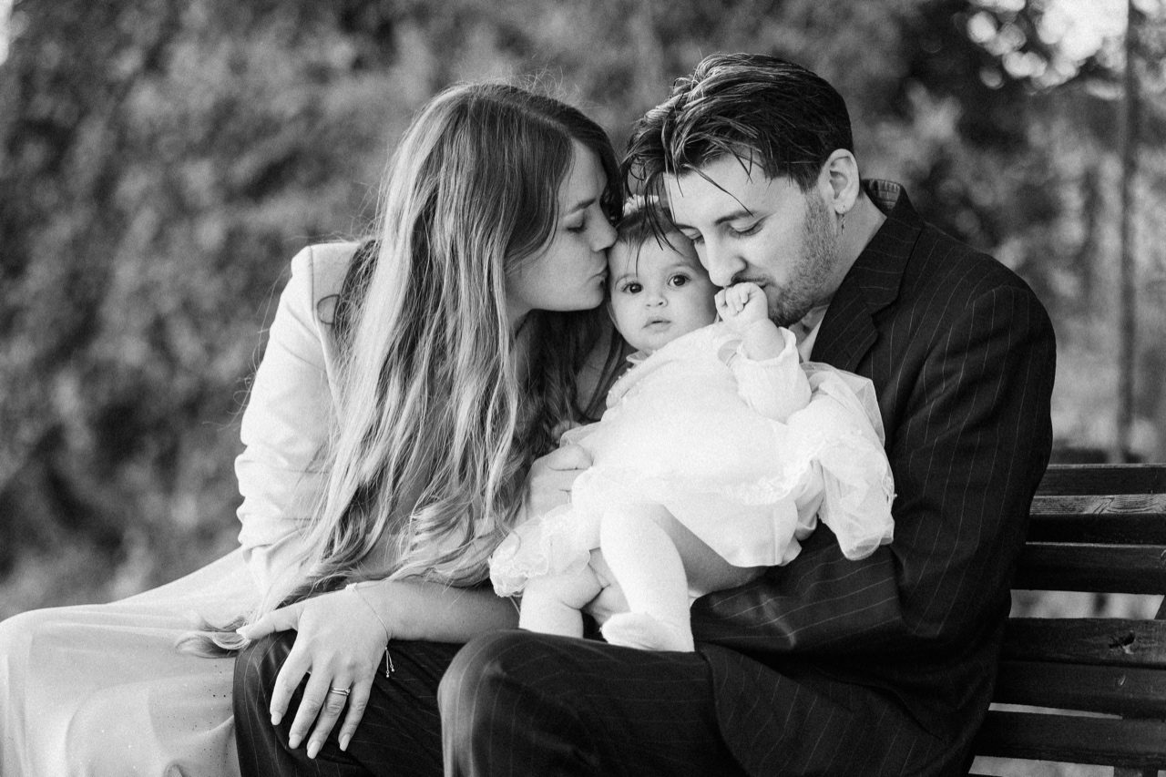Battesimo Roma — famiglia seduta sulla panca all'aperto, mamma e papà baciano la bambina, scatto reportage in bianco e nero