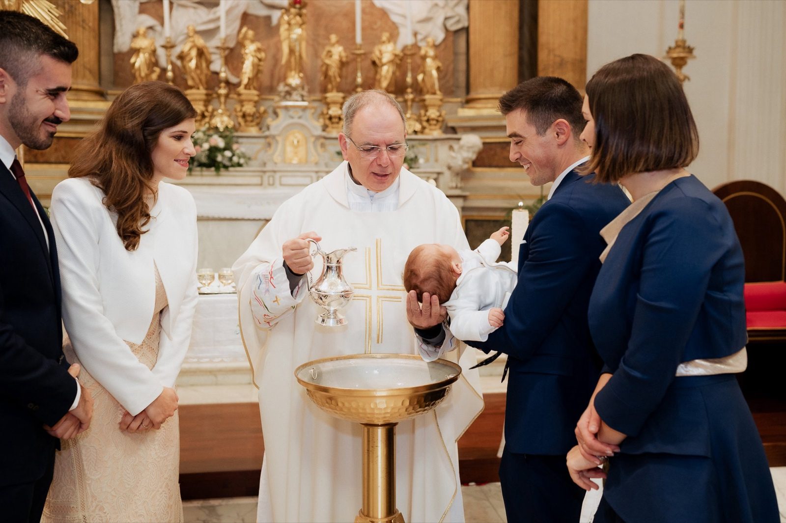 Cerimonia battesimo Roma — il sacerdote benedice il bambino con genitori e padrini al fonte battesimale durante il rito in chiesa