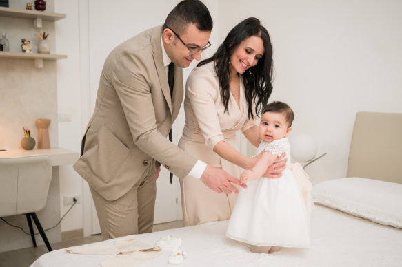 Preparativi battesimo Roma — genitori vestono la bambina sul letto, scatto reportage di Stefano Blejeru