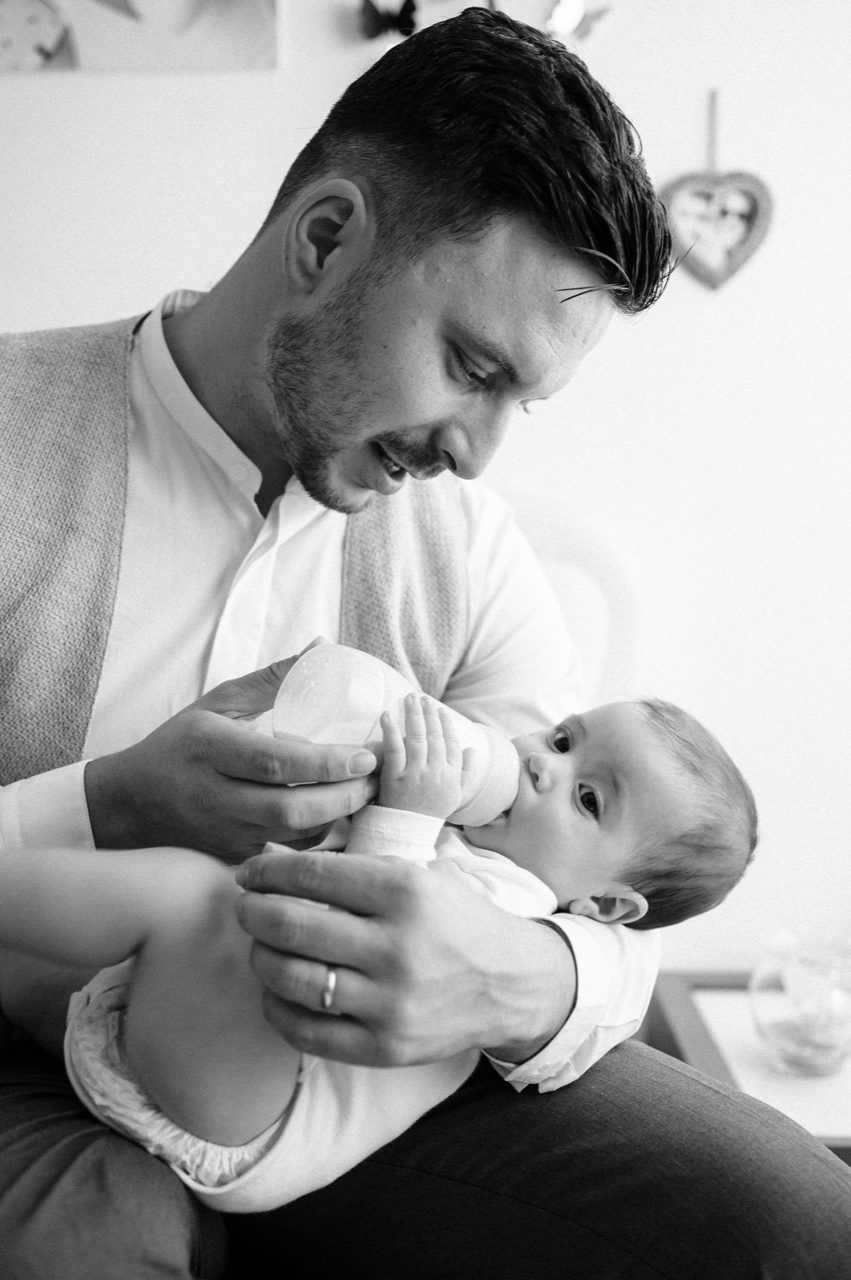 Preparativi battesimo Roma — papà dà il biberon al bambino in braccio, momento intimo in bianco e nero
